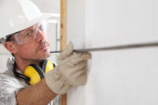 чоловік працівник вимірює кут стіни з квадратним носінням твердого капелюха, окулярів і навушників для захисту слуху, на внутрішньому будівельному майданчику
 - Фото, зображення