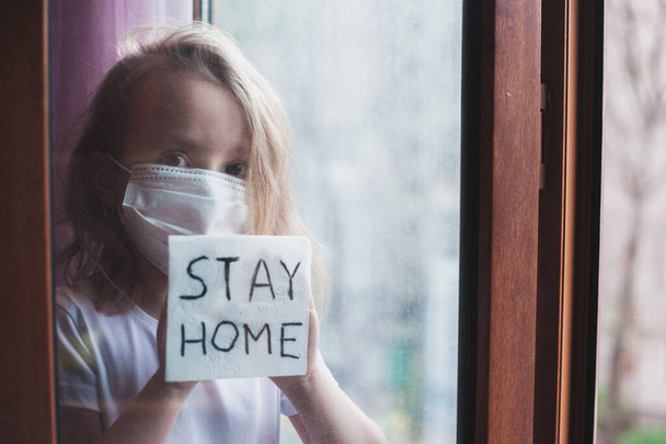 Zůstaňte doma v karanténě prevence pandemie koronaviru. Smutné dítě v ochranných lékařských maskách u oken a dívá se z okna. Pohled z ulice. Preventivní epidemie. Koncept koronaviru - Fotografie, Obrázek