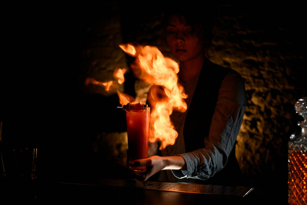 Woman bartender holding glass with cocktail sprinkles on it and masterfully make fire. - Fotografie, Obrázek