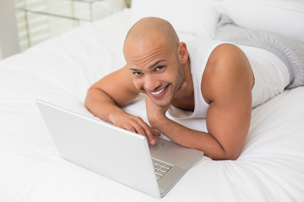 Smiling casual bald young man using laptop in bed - Photo, Image