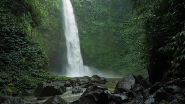 hämmästyttävä nungnung vesiputous hitaasti liikkuva putoava vesi osuu veden pintaan valtavia kiviä nähtävissä edessä runko rehevä vihreä lehdet liikkuu tuuli bali Indonesiassa
  - Materiaali, video