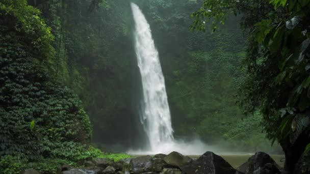 驚くべきヌンヌンの滝スローモーション落下水が水面を打つ緑豊かな葉のフレームの前に見えるいくつかの巨大な岩は風バリインドネシアから動いています - 映像、動画