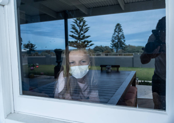 COVID-19, zamknięcie. Przygnębiona samotna dziewczynka z maską na twarzy patrząca przez okno podczas kwarantanny. Smutne chore dziecko w izolacji w domu. Epidemia Coronavirusu i zdrowie psychiczne dzieci. - Zdjęcie, obraz