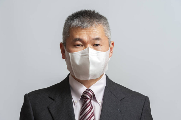 Vorderansicht eines asiatischen Mannes mittleren Alters mit Anzug und Krawatte, der eine weiße 3D-Einweg-Gesichtsmaske zum Schutz gegen das neuartige Coronavirus (SARS-CoV-2) und COVID-19 trägt. Vereinzelt auf weißem Hintergrund. Blick in die Kamera. - Foto, Bild