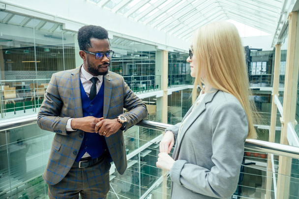 african american man in a white shirt in brown jacket posing talking to a business blonde caucasus woman in a gray suit in a modern office with glass windows shopping mall - Foto, afbeelding