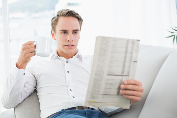 Relaxed man drinking coffee while reading newspaper on sofa - Фото, изображение