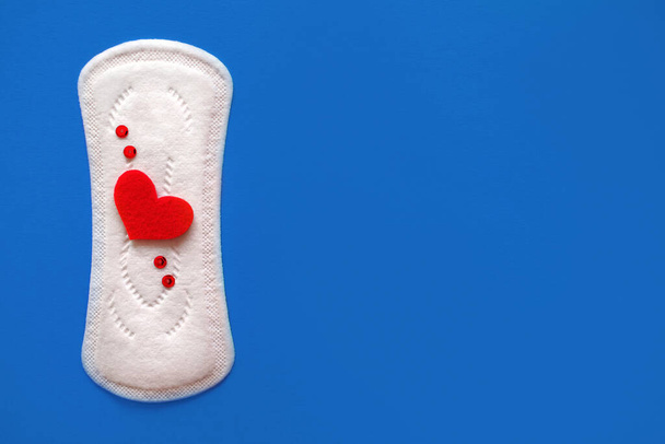 Menstruationspolster mit roten Funkeln in Herzform auf blauem Hintergrund, freier Raum für Text, das Konzept der Menstruation bei Frauen, ein Werkzeug zur Blutunterdrückung während des Menstruationszyklus - Foto, Bild