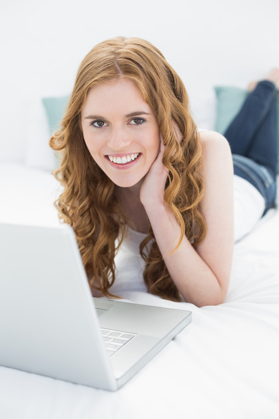 Portrait of smiling casual red-haired girl with laptop in bed - Foto, imagen