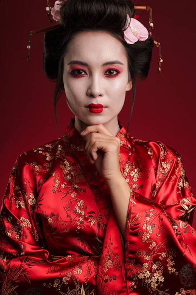 Image of attractive asian geisha woman in traditional japanese kimono touching her chin isolated over red background - Foto, Imagem