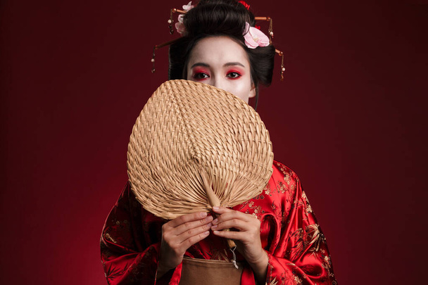 Imagem da jovem mulher gueixa em quimono tradicional japonês segurando ventilador de mão de madeira isolado sobre fundo vermelho
 - Foto, Imagem