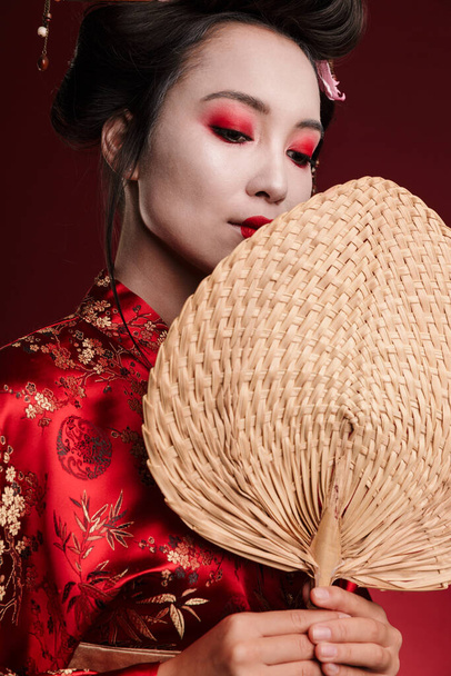 Image of young geisha woman in traditional japanese kimono holding wooden hand fan isolated over red background - Zdjęcie, obraz