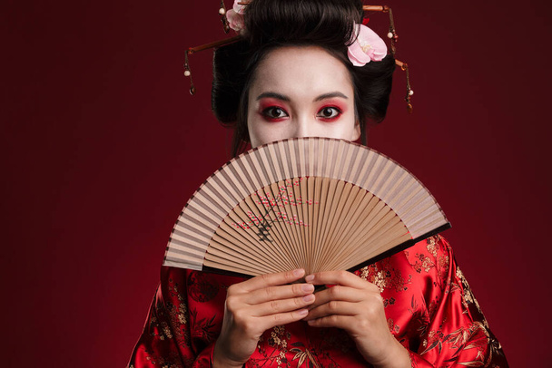 Image of young geisha woman in traditional japanese kimono holding wooden hand fan isolated over red background - Fotografie, Obrázek