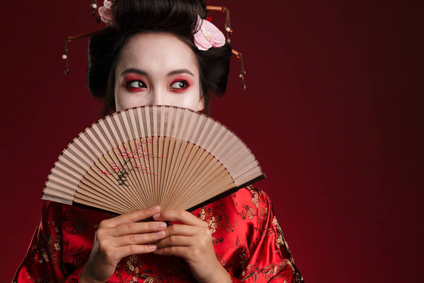 Image of young geisha woman in traditional japanese kimono holding wooden hand fan isolated over red background - Фото, изображение