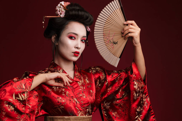 Image of young geisha woman in traditional japanese kimono holding wooden hand fan isolated over red background - Fotoğraf, Görsel
