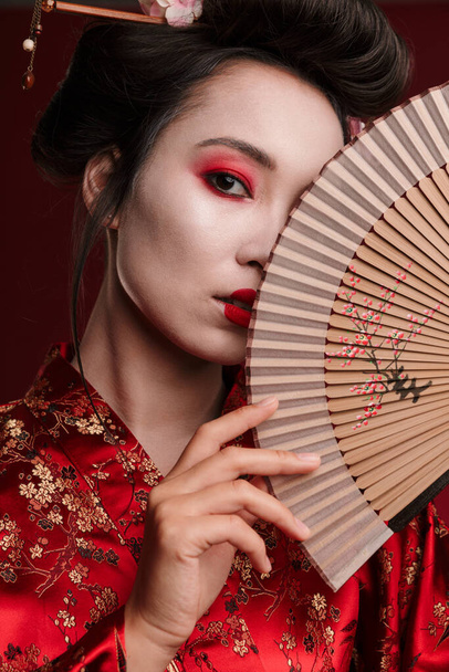 Image of young geisha woman in traditional japanese kimono holding wooden hand fan isolated over red background - Foto, immagini