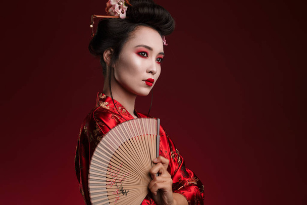 Image of young geisha woman in traditional japanese kimono holding wooden hand fan isolated over red background - Фото, изображение