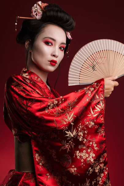 Image of young geisha woman in traditional japanese kimono holding wooden hand fan isolated over red background - Photo, image