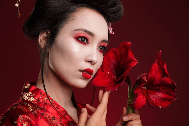 Image of beautiful geisha woman in traditional japanese kimono holding flowers isolated over red background - Foto, Bild