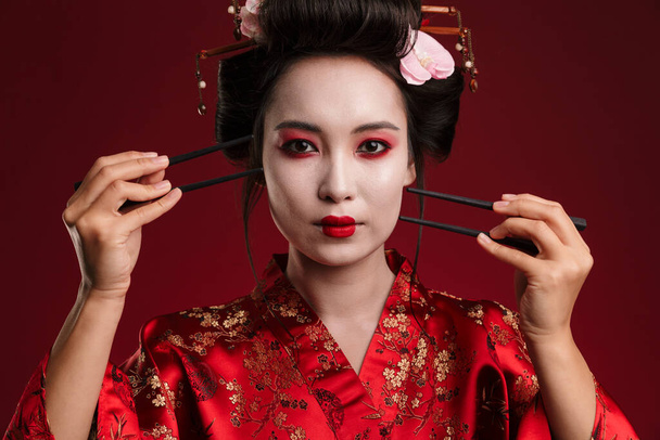 Image of beautiful geisha woman in traditional japanese kimono holding chopsticks isolated over red background - Photo, image