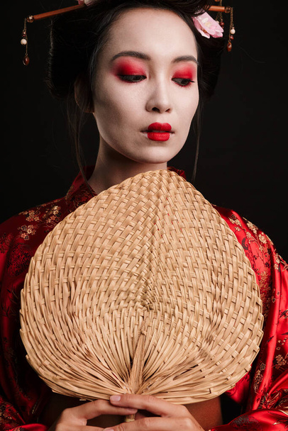 Image of young geisha woman in traditional japanese kimono holding wooden hand fan isolated over black background - Fotografie, Obrázek