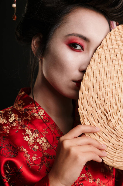 Image of young geisha woman in traditional japanese kimono holding wooden hand fan isolated over black background - Photo, image