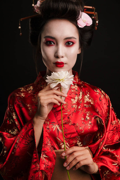 Image of beautiful geisha woman in traditional japanese kimono holding flower isolated over black background - Zdjęcie, obraz