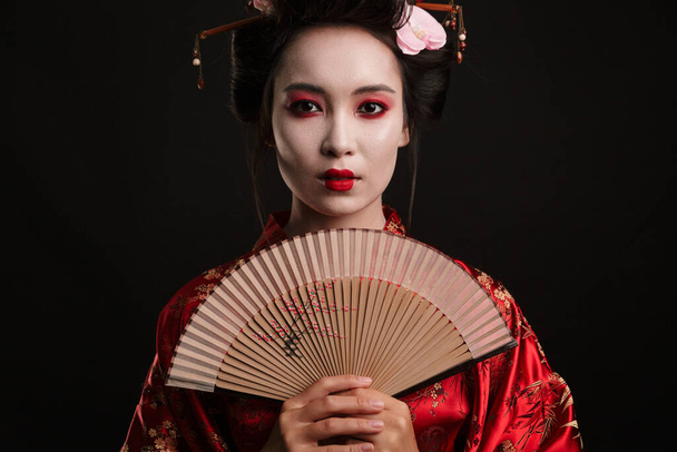 Image of young geisha woman in traditional japanese kimono holding wooden hand fan isolated over black background - Foto, Bild