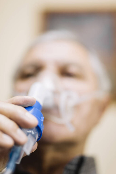 Senior man using medical equipment for inhalation with respiratory mask, nebulizer in the room - Фото, изображение