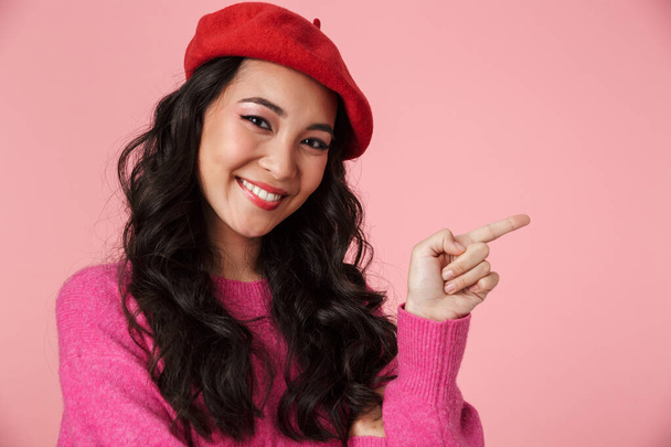 Image of attractive beautiful asian girl wearing beret laughing and pointing finger at copyspace isolated over pink background - Foto, immagini