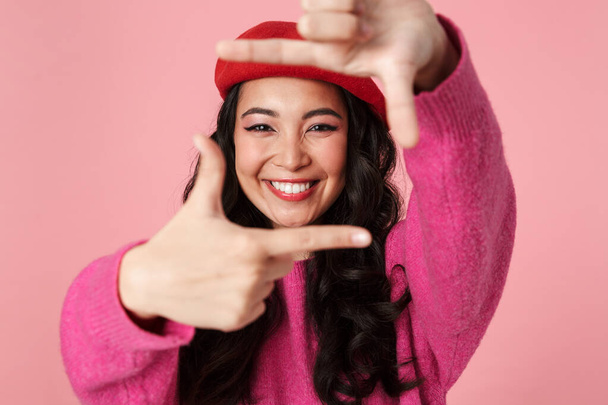 Image of content beautiful asian girl wearing beret smiling and showing photo frame with her fingers isolated over pink background - Foto, immagini
