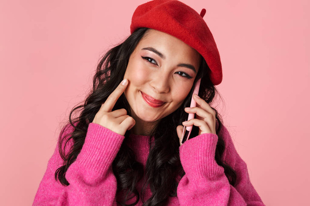 Image of optimistic beautiful asian girl wearing beret smiling and talking on smartphone isolated over pink background - Foto, immagini