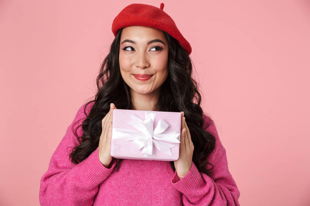 Obrázek vzrušené krásné asijské dívky s dlouhými tmavými vlasy na sobě baret s úsměvem a držení dárek box izolované přes růžové pozadí - Fotografie, Obrázek
