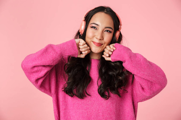 Image of optimistic beautiful asian girl with long dark hair smiling and listening to music with headphones isolated over pink background - Φωτογραφία, εικόνα