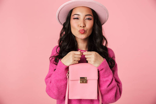Изображение прекрасной красивой азиатской девушки с длинными темными волосами в шляпе улыбаясь и держа мешок изолированы на розовом фоне
 - Фото, изображение