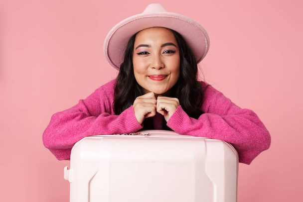 Şapka takan, gülümseyen ve elinde pembe arka planda izole edilmiş seyahat çantası tutan güzel Asyalı bir kız resmi. - Fotoğraf, Görsel