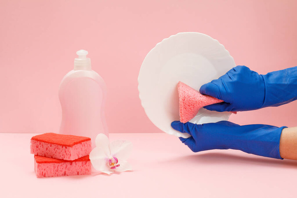 Les mains des femmes en gants de protection en caoutchouc avec une plaque blanche et une éponge, une bouteille de liquide vaisselle, éponges sur un fond rose. Concept de lavage et de nettoyage
. - Photo, image