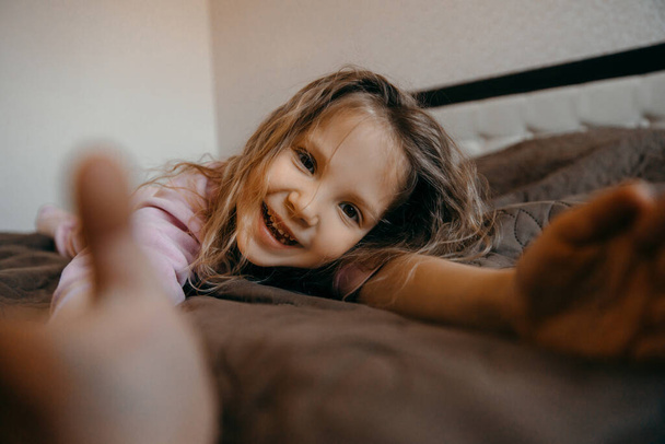 Bambino ragazza divertirsi pigiama party durante Covid-19 quarantena a casa. Divertente bambina sdraiata sul letto e sorridente
.   - Foto, immagini