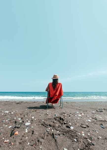 Girl in hat sitting on a lounger with orange towel on the shore of the beach - Foto, afbeelding