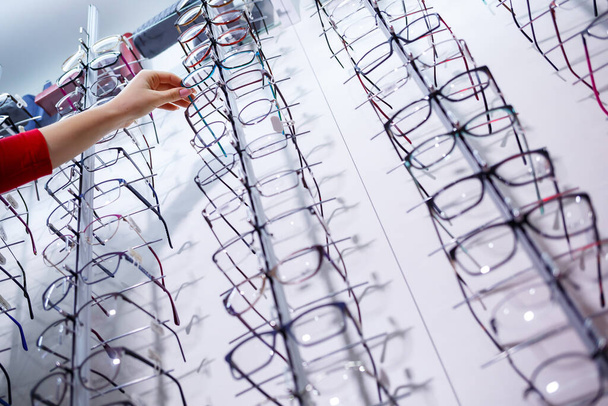 Row of glasses at an opticians. Eyeglasses shop. Stand with glasses in the store of optics. Woman's hand shows glasses. Presenting spectacles. Closeup - Photo, Image
