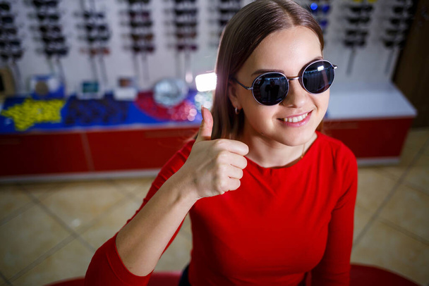 Όμορφη νεαρή κοπέλα επιλέγει γυαλιά ηλίου σε ένα κατάστημα οπτικών. - Φωτογραφία, εικόνα