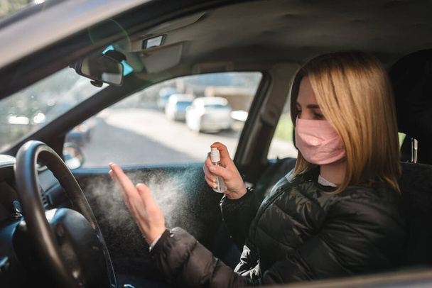 Spraying antibacterial sanitizer spray on hand in car, infection control concept. Sanitizer to prevent Coronavirus, COVID-19, flu. Spray bottle. Woman wearing in medical protective mask driving a car. - Фото, изображение