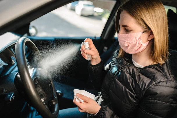 Spraying antibacterial sanitizer spray on steering wheel, disinfection car, infection control concept. Prevent Coronavirus, COVID-19, flu. Woman wearing in medical protective mask driving a car. - Foto, afbeelding