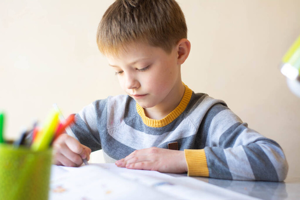 Poika tekee läksynsä. Lapsi oppii kauko-ohjatusti. Poika istuu pöydän ääressä ja kirjoittaa. Online-oppiminen
 - Valokuva, kuva