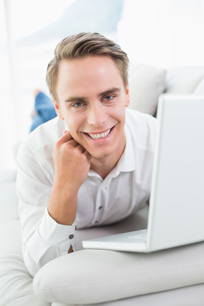 Smiling young man with laptop lying on sofa - Zdjęcie, obraz