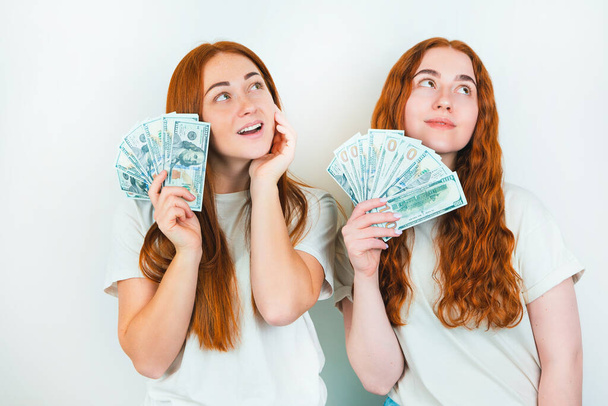 two redheaded young women both holding cash like playing cards looking thoughful standing on isolated white backgroung, money earning concept. - Foto, Imagen