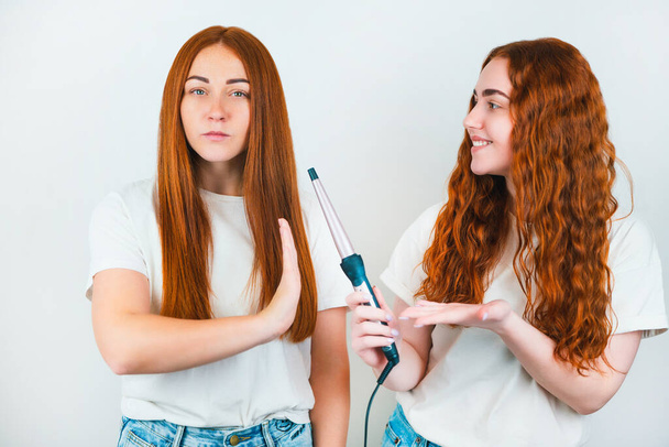 two redheaded young women standing on isolated white backgroung, one wants to curl her friend's hair with styler, but she refuses ,style and beauty concept. - Photo, image