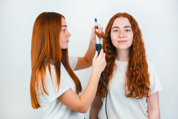 two redheaded young women standing on isolated white backgroung, one curls her friend's hair with styler, both look satisfied ,style and beauty concept. - Photo, Image
