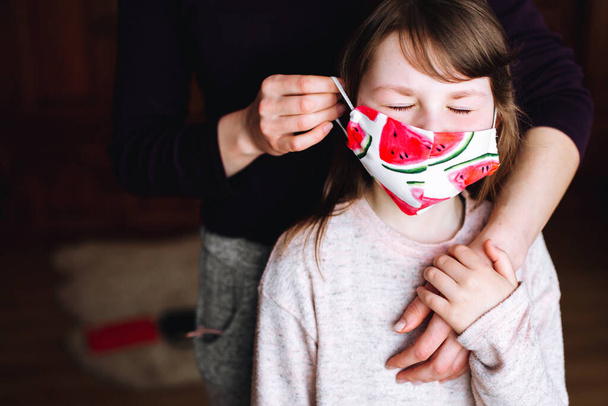 Matka zakłada córce maskę z jasnym odciskiem arbuza, żeby ochronić ją przed wirusem. Pandemia koronawirusa. Zostań w domu, by powstrzymać rozprzestrzenianie się korony.. - Zdjęcie, obraz