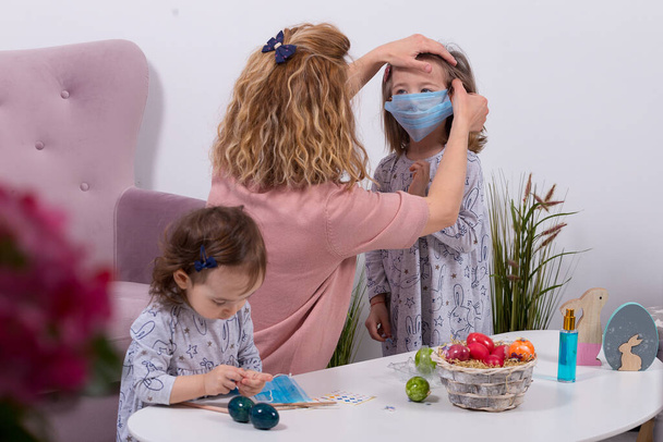 Matka a dcery malují vejce. Šťastná rodina se připravuje na Velikonoce. Matka nosila masku své dcery proti koronaviru. Karanténa doma. - Fotografie, Obrázek