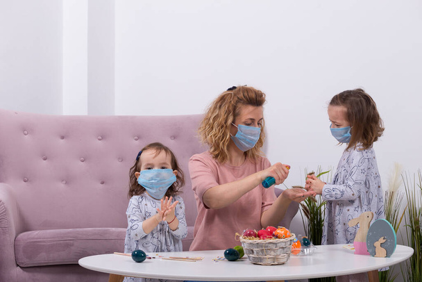 Matka a dcery malují vejce. Šťastná rodina se připravuje na Velikonoce. Matka nosila masku své dcery proti koronaviru. Karanténa doma. - Fotografie, Obrázek